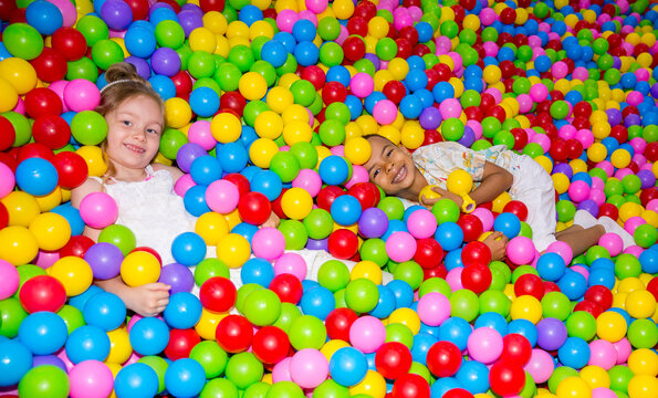 Happy girl playing in ball pit on birthday party in kids amusement park and indoor play center. Child playing with colorful balls in playground ball pool.