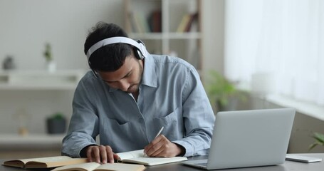 Focused serious Indian student guy in headphones watching online learning conference on laptop, talking on video call to teacher, studying, writing notes, speaking - Powered by Adobe