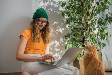 A female freelancer is working on a laptop sitting on the floor in a room, a red cat is sitting...