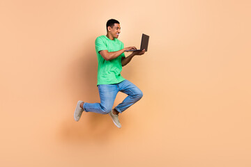 Full length photo of good mood man wear stylish t-shirt jeans jumping check fast internet on laptop isolated on beige color background