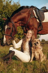 A young beautiful woman jockey with her dog sits in a meadow near her horse at sunset.