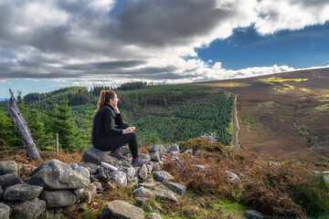 Adult woman sitting on a rock, drinking tea or coffee from thermos bottle and looking at view....