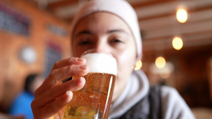 Woman drinking draft beer at restaurant during cold season. Close-up face of person drinks refreshing alcoholic beverage - obrazy, fototapety, plakaty