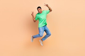 Fototapeta na wymiar Full size photo of good mood cheerful guy dressed green t-shirt denim pants flying showing v-sign isolated on beige color background