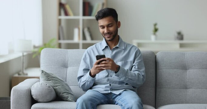 Serious young Indian man chatting on mobile phone, reading message on smartphone at home, sitting on sofa, getting happy, positive, turning look away, smiling, thinking on answer