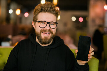 Satisfied man with dark beard holding smartphone and showing his favorite mobile app Male...
