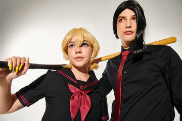 couple of cosplayers in wigs and school uniform with baseball bats looking away on grey, subculture