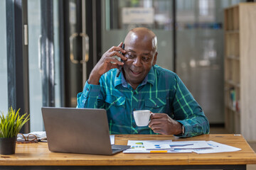 Senior black man talking on phone with attention tracking data on laptop screen. Sip coffee while talking on the phone.