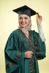 A female muslim lady wearing hijab and convocation attire smiling, isolated on yellow background