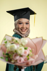 A female muslim lady wearing hijab and convocation attire smiling, isolated on yellow background