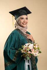 A female muslim lady wearing hijab and convocation attire smiling, isolated on brown background