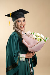A female muslim lady wearing hijab and convocation attire smiling, isolated on brown background