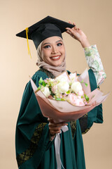 A female muslim lady wearing hijab and convocation attire smiling, isolated on brown background