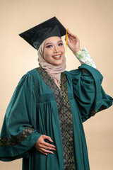 A female muslim lady wearing hijab and convocation attire smiling, isolated on brown background