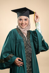 A female muslim lady wearing hijab and convocation attire smiling, isolated on brown background