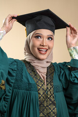 A female muslim lady wearing hijab and convocation attire smiling, isolated on brown background