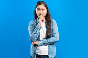 Portrait of an attractive Indian girl showing shh sign with forefinger against blue backdrop