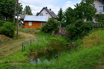 A close up on a small pond or river flowing next to a dirt path located near some urban settlements and lush trees and shrubs seen on a cloudy summer day on a Polish countryside during a hike