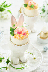 Stuffed or deviled eggs with yolk, shrimp, pea microgreens with paprika in rabbit-shaped stand for easter table decorate fresh cherry or apple blossoms on light background. Traditional dish for Easter