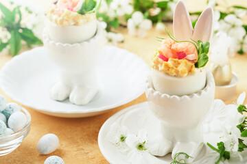 Stuffed or deviled eggs with yolk, shrimp, pea microgreens with paprika in rabbit-shaped stand for easter table decorate fresh cherry or apple blossoms on light background. Traditional dish for Easter