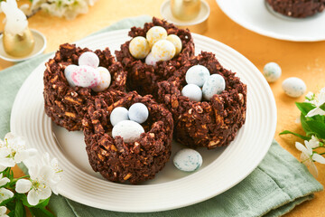 Easter chocolate nest cake with mini chocolate candy eggs with blossoming cherry or apple flowers on yellow background table. Creative recipe for Easter table with holiday decorations. Top view.