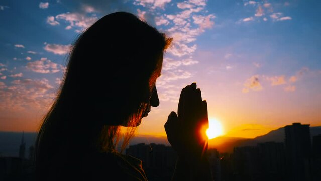 Close up: woman silhouette is folding hands and praying against the sunrise or sunset sky over the city: side view, sun lens flares. Religion, prayer gesture, hope, spiritual and nature concept