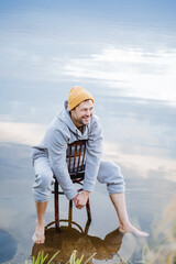 Guy smiling, laughter and joy, man sitting on chair in water, bare feet hipster resting on pond.