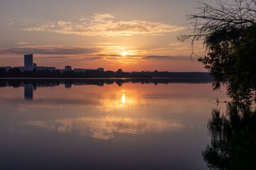 Scenic blue sunrise sun on cloudy sky reflecting in calm lake water. Peaceful morning in city park by the river