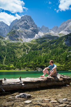 Ein Wanderer sitzt auf einem Baumstamm im Wasser und beweundert das Bergpanorma der Mieminger Kette am Seebensee.
