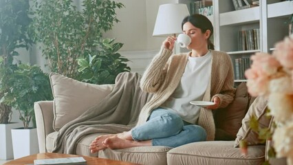 Relaxed woman drinking coffee or herbal tea and feeling carefree and refreshed while relaxing on the couch at home. Break time. Girl enjoying comfortable relaxing sitting on couch in the morning. 4K - Powered by Adobe