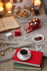 Bowl of cookies, cup of tea, dry oranges, pine cones, books, reading glasses, small presents, various Christmas decorations and lit candles on the table. Cozy Christmas hygge. Selective focus.
