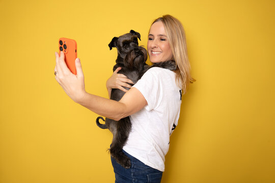 Friendship concept. Happy european woman taking selfie with her cute schnauzer dog, looking and smiling at camera over yellow studio background wall