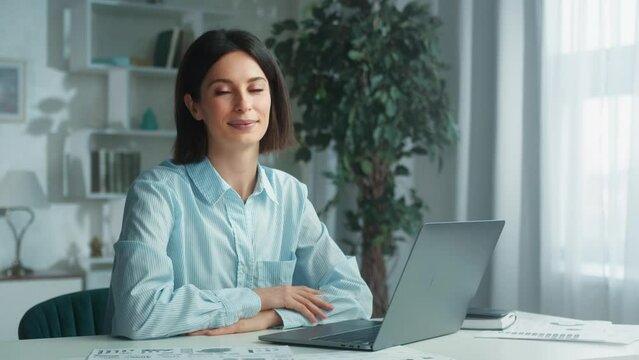 A young brunette girl looks at the camera and smiles at home at her workplace in the office. Female freelancer, successful motivated employee, secretary portrait, modern wireless tech usage concept. 4
