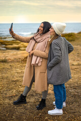Two young lesbian women enjoy looking at a newly taken photo in selfie by the winter sea. LGBTQ couple enjoys technology on vacation, calling prori friends in video call with smartphone