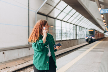 Young refugee redhead woman crying and waiting train on station platform, she lost or late, unhappy girl talking on smart phone. Railroad transport concept