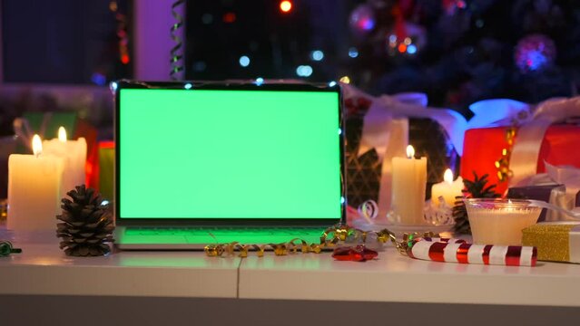 Laptop with green chromatic screen close-up, burning candle, gift boxes, tinsel, bokeh and xmas tree. Christmas background. New Year holidays concept.