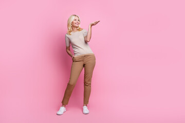 Full length photo of good mood woman wear trendy clothes brown trousers look empty space product on arm isolated on pink color background