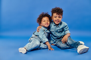 adorable african american siblings in stylish denim clothes sitting and hugging on blue background