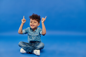 joyful and curly african american preschooler boy in denim clothes sitting pointing up on blue