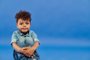 positive african american preschooler boy in stylish denim clothes sitting on blue background