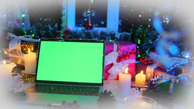 Open laptop with chromakey on screen close up on desk among Christmas tree decor, burning candles, tinsel, pine cones and flickering colored garlands, creating xmas mood, during New Year time.