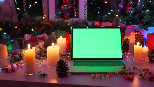 Festive winter holiday decor laptop with green screen, candles and traditional Christmas tree in tinsel and flickering garlands, close up. Celebration Happy New Year background. Advertising platform.