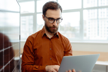 Handsome serious bearded man, programmer, developer using laptop computer working online in office,...