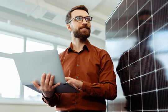 Handsome smiling man, engineer, developer holding laptop computer planning startup working with solar panel in modern office. Innovation, renewable energy concept  