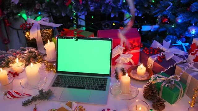 Close up, laptop computer with green screen near warm candlelight, gifts, presents, cones, flashing garland on Christmas celebration background. New Year holidays.