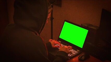 Female hacker sits in front of green laptop screen in dark room wearing hood. Cybercrime concept.