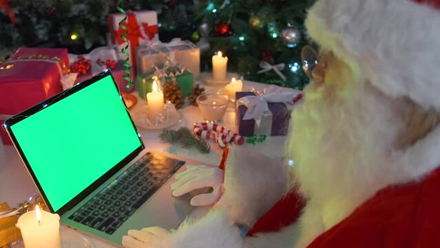 Santa Claus adjusts his glasses, reads letters from children at email, looks at green laptop screen, among Christmas decorations, candles and gift boxes, next to Christmas tree and twinkling garlands.