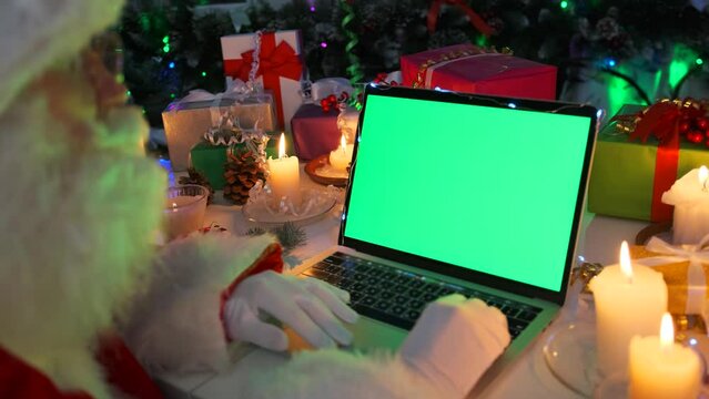 Santa Klaus with beard and glasses sitting at laptop with chromakey, using touchpad, reading emails with children wishes, among wrapped gifts boxes, burning candles, cones, tinsel and garlands.