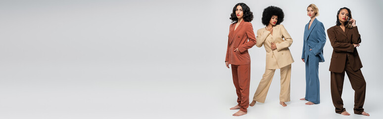 multiracial barefoot girlfriends in trendy multicolored suits posing on grey, horizontal banner