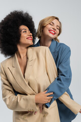 delighted woman embracing african american girlfriend while posing in colorful suits on grey